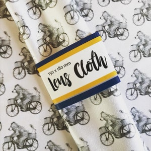 May include: White fabric with a repeating pattern of black and white bears riding bicycles. The fabric has a yellow and blue label that says "Lens Cloth" and "150 x 180 mm".