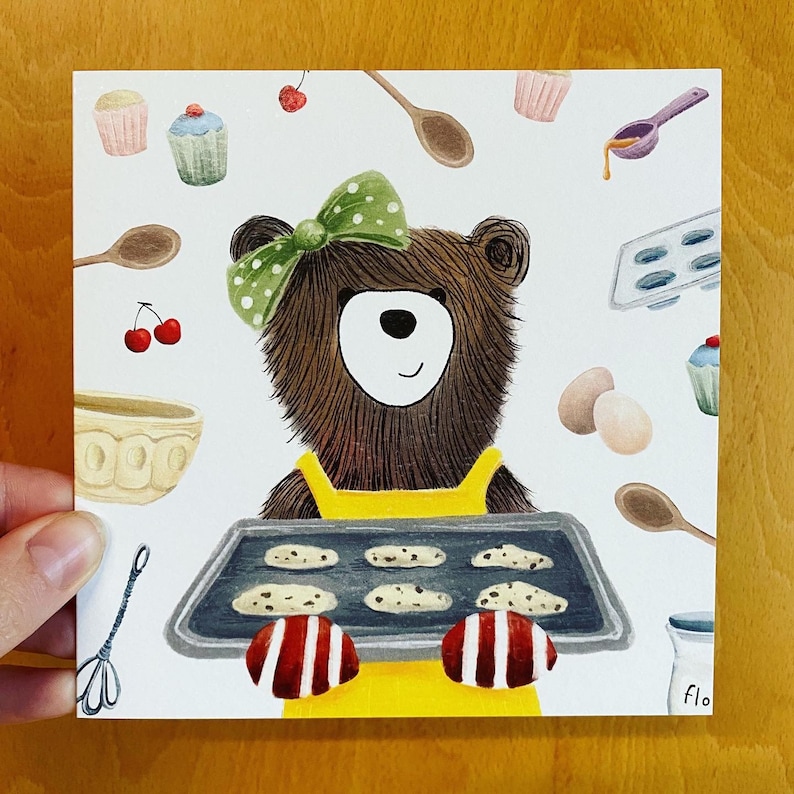 Puede incluir: Un oso marr&oacute;n con un delantal amarillo y un lazo verde est&aacute; horneando galletas. El oso sostiene una bandeja para hornear con seis galletas. La bandeja para hornear est&aacute; sobre un fondo blanco con otros utensilios para hornear.