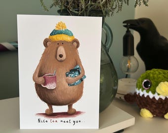 Puede incluir: Tarjeta de felicitación con un oso pardo con un gorro amarillo y turquesa, sosteniendo una taza y libros. La tarjeta dice "Nice tea meet you..". La tarjeta está sobre una superficie blanca.