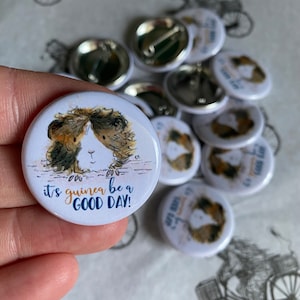 May include: A white button with a cartoon guinea pig on it. The guinea pig is brown and white with black eyes. The text on the button reads "It's guinea be a good day!"