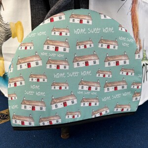 May include: A teal blue tea cosy with a repeating pattern of white houses with red doors and the text "home sweet home".