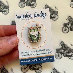 May include: A wooden badge featuring a green creature with a blue scarf and flowers on its head. The badge is on a white card with the text "Wooden Badge by Catherine Redgate" and "This badge was made in the UK using responsibly sourced maple veneer MDF www.catherine-redgate.co.uk".