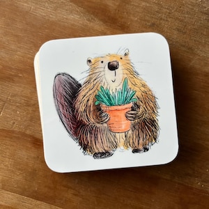 May include: Square white coaster with a cartoon beaver holding a small orange flower pot with green plants. The beaver has brown and tan fur, a dark brown tail, and a black nose. The coaster has rounded corners and is set against a wooden background.