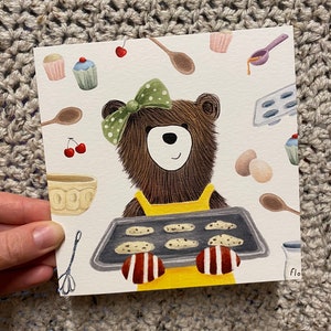 Puede incluir: Un oso marr&oacute;n con un delantal amarillo y un lazo verde sostiene una bandeja para hornear con galletas. La bandeja para hornear tiene seis galletas. El oso est&aacute; rodeado de varios utensilios de reposter&iacute;a, como un batidor, una cuchara de madera y un taz&oacute;n.