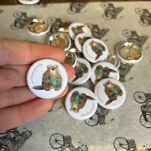 May include: A selection of white, circular badges featuring a cartoon beaver. The beaver is depicted wearing glasses and holding a green mug, with the text "just beavering away..." in a cursive font. The badges are on a patterned surface.