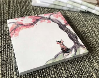 Puede incluir: Un bloc de notas blanco con una ilustración de acuarela de un zorro sentado debajo de un cerezo en flor rosa. El bloc de notas tiene forma cuadrada y es perfecto para tomar notas o escribir ideas.