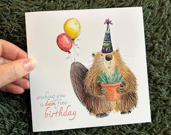 May include: A birthday card featuring a cartoon beaver wearing a party hat and holding a potted plant. Two balloons float above the beaver. The card reads "wishing you a dam fine birthday" in red and blue script.