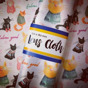 May include: White fabric with a repeating pattern of cats wearing sweaters. The fabric has the text "feline good" and "Lens Cloth" printed on it. The fabric measures 150 x 180 mm.