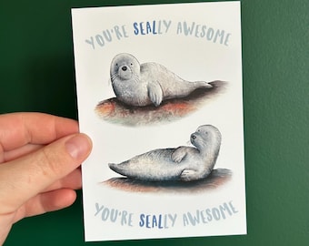 May include: A white greeting card with two illustrations of seals. The text "YOU'RE SEALLY AWESOME" is written in blue above and below the seals. The seals are gray and lying on a brown surface.