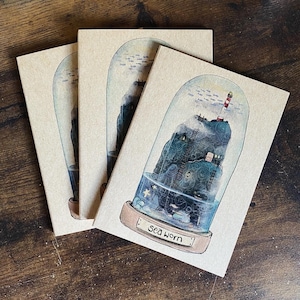 Op de afbeelding: Drie bruine papieren notitieboekjes met een aquarel-illustratie van een vuurtoren op een rotsachtig eiland in een glazen koepel. De tekst "Sea Worn" staat gedrukt op de voorkant van het notitieboekje.