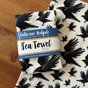 May include: White tea towel featuring a black raven design. The tea towel measures 72cm x 46cm. The label reads "Tea Towel" and "Catherine Redgate". Made in the UK.