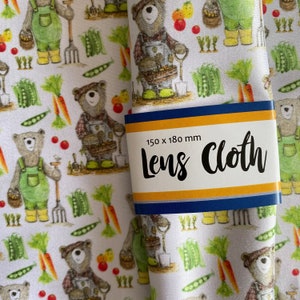 May include: White fabric with a repeating pattern of brown bears wearing overalls and holding gardening tools. The bears are surrounded by carrots, peas, and tomatoes. The fabric has a label that reads "Lens Cloth 150 x 180 mm".