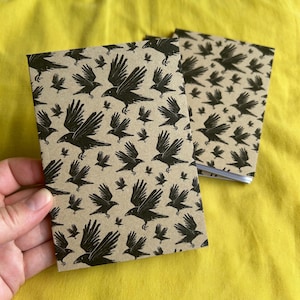 May include: Two small notebooks with a repeating pattern of black crows on a tan background. The crows are depicted in various sizes and poses, with some in flight. The notebooks are held against a yellow background.
