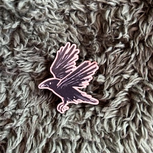 May include: A black and pink sticker of a crow in flight. The crow is illustrated with detailed wing feathers and a curved beak. The sticker has a pink border and is set against a textured, grey background.