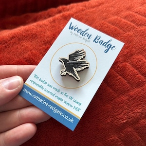 May include: A wooden raven badge on a white card. The card reads "Wooden Badge by Catherine Redgate". The badge is black with a natural wood edge. Text on the card states "This badge was made in the UK using responsibly sourced maple veneer MDF".
