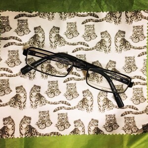 May include: A white microfiber cleaning cloth with a repeating pattern of black and white snow leopards. A pair of black rectangular glasses is lying on top of the cloth.