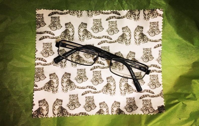 May include: A white microfiber cleaning cloth with a repeating pattern of black and white snow leopards. A pair of black rectangular glasses is lying on top of the cloth.