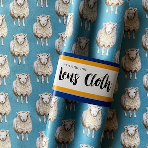 Peut inclure: Un tissu bleu avec un motif de moutons blancs. Le tissu est plié et porte une étiquette indiquant "150 x 180 mm Lens Cloth".