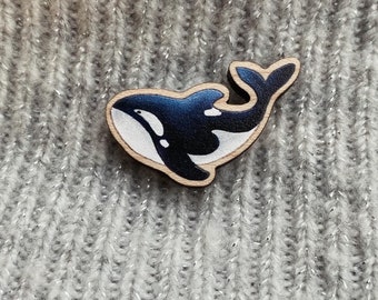 May include: A wooden pin with a black and white painted orca whale design. The orca is swimming to the right. The pin is on a gray knitted fabric background.