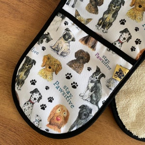 May include: White oven mitt with a black border and a pattern of dogs and paw prints. The text "stay pawsitive" is repeated on the mitt.