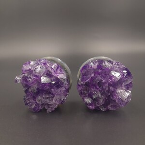 Könnte beinhalten: Ein Paar Amethyst-Kristall-Stecker, jeweils mit einer silbernen Rückseite. Die Kristalle sind tiefviolett und haben eine raue, natürliche Textur.