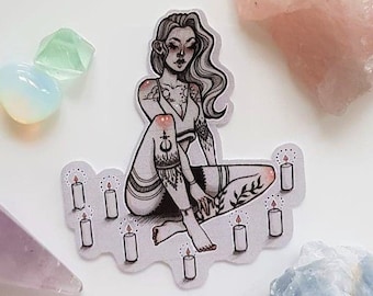 May include: A sticker featuring a seated woman with tattoos, surrounded by lit candles. The illustration is in black and white with intricate details. Various crystals in shades of pink, blue, and green are scattered around the sticker on a white background.
