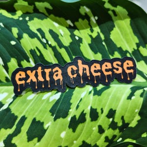 Puede incluir: Una pegatina negra con las palabras "extra cheese" en naranja, con letras goteando. La pegatina está colocada sobre una gran hoja verde con un patrón moteado.