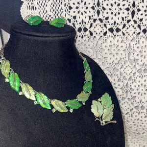 Könnte beinhalten: Ein grünes und silbernes Blatt-Halsband und Ohrringe mit einer passenden Brosche. Die Halskette ist aus Silber mit grünen Emailleblättern und klaren Strasssteinen. Die Ohrringe und die Brosche sind ebenfalls aus Silber mit grünen Emailleblättern.