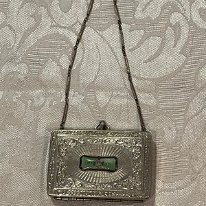 May include: A silver hinged compact with an ornate design and a green enamel panel with a floral design. The compact is attached to a silver chain.