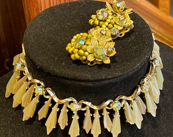 May include: A gold tone necklace with a row of iridescent rhinestones and a dangling row of cream colored, leaf-shaped pendants. A matching pair of earrings with green and gold tone flowers and iridescent rhinestones.