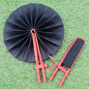 May include: Two black fabric folding fans with red leather trim and wooden handles. The fans are closed and lying on a green grassy surface.