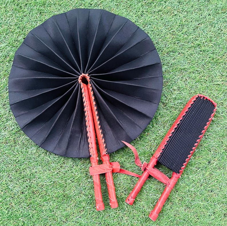 May include: Two black fabric folding fans with red leather trim and wooden handles. The fans are closed and lying on a green grassy surface.