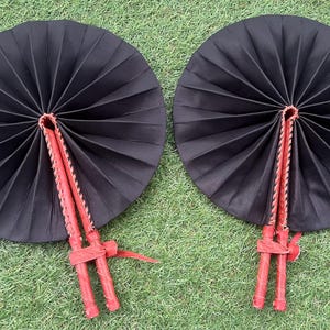 May include: Two black folding fans with red wooden handles. The fans are closed and have a pleated design.