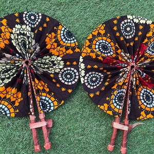 Może przedstawiać: Dwa wachlarze ręczne z brązowej tkaniny z pomarańczowymi, białymi i niebieskimi wzorami geometrycznymi. Wachlarze są złożone i mają drewniane uchwyty.