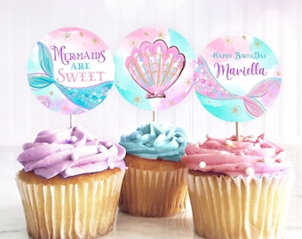 May include: Three cupcakes decorated with pink, purple, and blue frosting. Each cupcake has a round, colorful paper topper with a mermaid tail, seashell, and the text "Mermaids are Sweet" and "Happy Birthday Mariella".