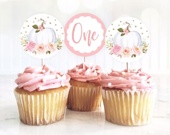 May include: Three pink and white cupcakes with white frosting and gold sprinkles. Each cupcake has a round topper with a white pumpkin, pink flowers, and gold glitter. The middle topper says "One" in pink script.