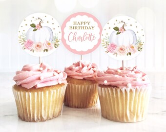 May include: Three pink and white cupcakes with white frosting and gold sprinkles. Each cupcake has a round topper with a white pumpkin, pink flowers, and gold glitter. The center topper says "Happy Birthday Charlotte".
