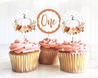 May include: Three cupcakes with pink frosting and white sprinkles topped with round paper cupcake toppers featuring a white pumpkin with orange and brown floral accents, gold glitter, and the word "One" in brown text.