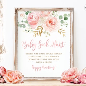 May include: A printable sign with a pink floral design and gold accents. The sign says "Baby Sock Hunt" and includes instructions for a baby shower game. There are baby socks hidden throughout the shower, and whoever finds the most wins a prize. The sign also says "Happy Hunting!"