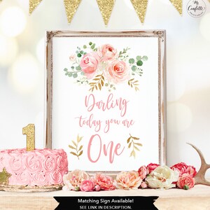 May include: A framed print with floral art and the text "Darling today you are One." A pink cake with a gold "1" candle sits next to the print. Gold glitter banner hangs above. The words "Matching Sign Available! SEE LINK IN DESCRIPTION" are at the bottom.