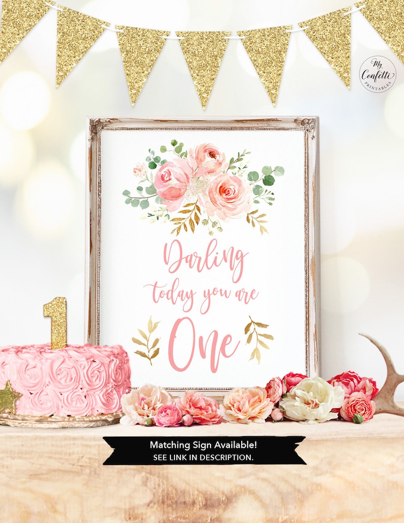 May include: A framed print with floral art and the text "Darling today you are One." A pink cake with a gold "1" candle sits next to the print. Gold glitter banner hangs above. The words "Matching Sign Available! SEE LINK IN DESCRIPTION" are at the bottom.