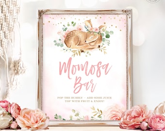 May include: A printable sign for a mimosa bar with a pink background and gold glitter. The sign features a watercolor illustration of a fawn with a floral crown, surrounded by greenery and pink flowers. The text on the sign reads "Momosa Bar" and "Pop the bubbly - add some juice top with fruit & enjoy!"