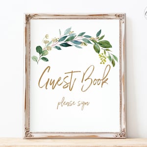 Puede incluir: Un cartel imprimible con fondo blanco y marco dorado. El cartel presenta una ilustración de acuarela de una guirnalda de hojas verdes con detalles dorados. El texto "Guest Book" está escrito en letra dorada de estilo cursivo y el texto "please sign" está escrito en una letra dorada más pequeña.