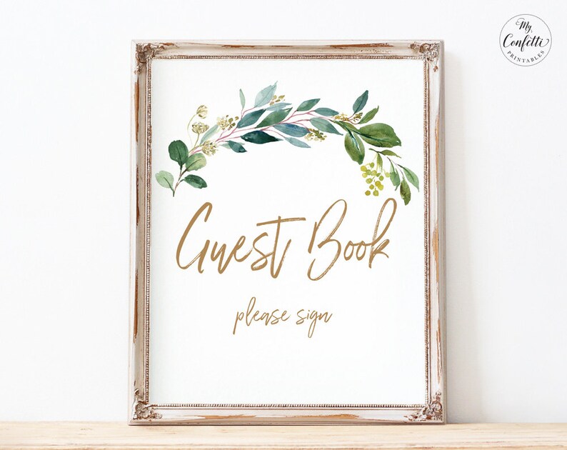 May include: A printable sign with a white background and a gold frame. The sign features a watercolor illustration of a green leafy garland with gold accents. The text "Guest Book" is written in gold script font and the text "please sign" is written in a smaller gold font.