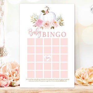 May include: A printable baby shower bingo card with a pink and white floral design featuring a white pumpkin with pink flowers. The card has a free space in the center and 24 other squares to fill in with gifts the new mom will receive.