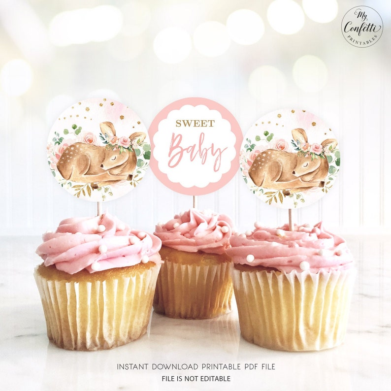May include: Three pink frosted cupcakes with gold glitter cupcake toppers featuring a sleeping fawn with a floral crown and the text "Sweet Baby".