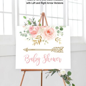 May include: A white sign with a pink floral design and a gold arrow pointing to the right. The text "Baby Shower" is written in pink cursive lettering below the arrow. The sign is on a wooden easel.