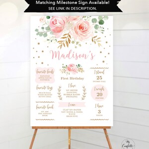 May include: A milestone sign for a first birthday, featuring a floral design with pink roses and gold accents. The sign includes the name "Madison's", favorite foods, toys, and book, along with height (25 inches), weight (20 pounds), and number of teeth (3).