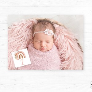 May include: A baby girl wearing a white headband with flowers is lying on a pink fluffy blanket. A white card with a rainbow and the number "2" is in the foreground.