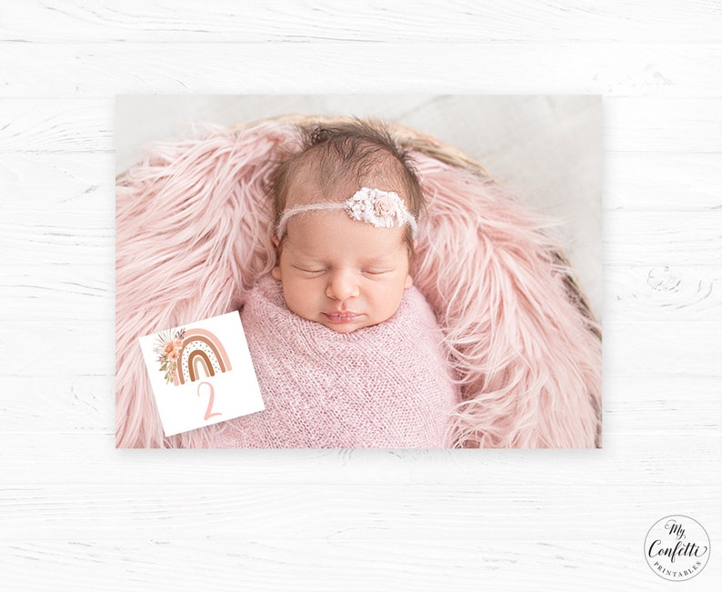 May include: A baby girl wearing a white headband with flowers is lying on a pink fluffy blanket. A white card with a rainbow and the number "2" is in the foreground.